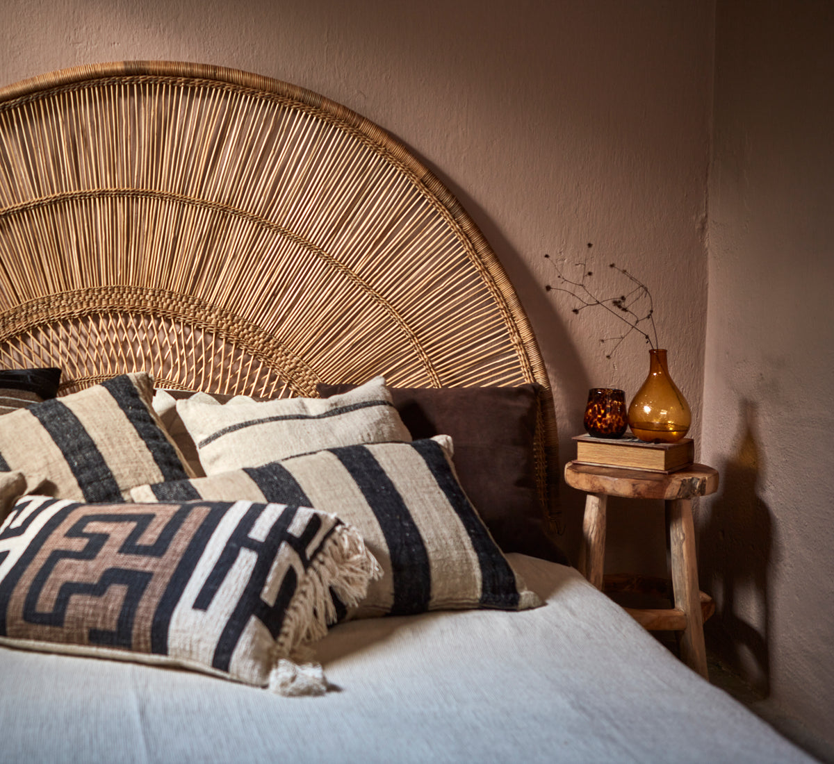 Bed with wicker headboard and decorative cushions, next to a small wooden table with a bottle and books.