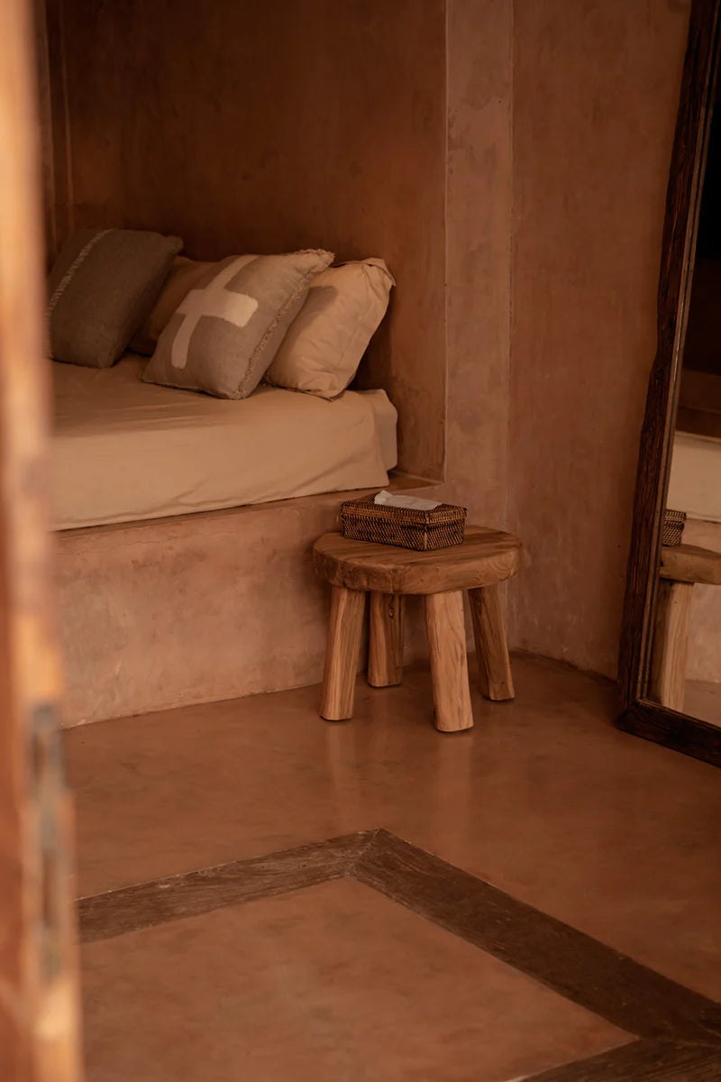 Small bedroom with a bed, wooden stool, and basket in a warm toned room with natural lined cushions