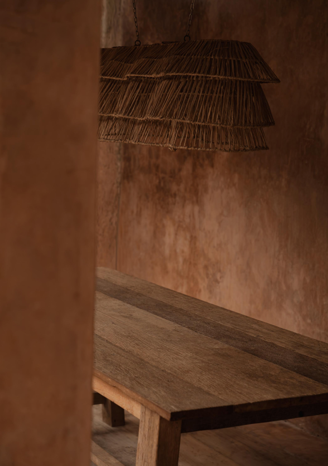 Rustic wooden table with a wicker rattan long statement pendant light against a textured wall.