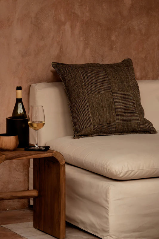 Beige sofa with a black and brown linen cushion, wine bottle, and glass on a wooden side table against a textured wall.