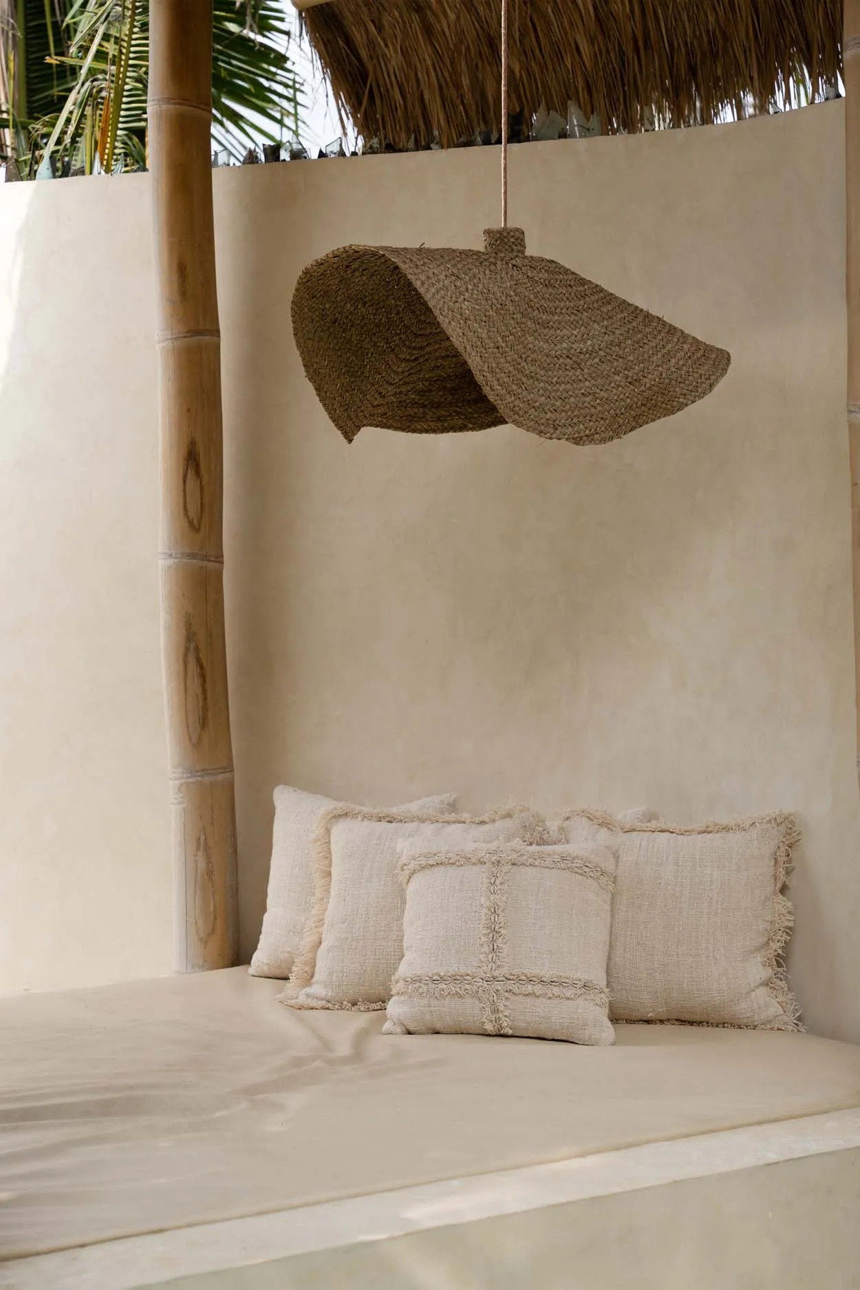 Beige interior with pillows on a bench and a woven lampshade hanging from a thatched roof.
