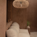 A woven natural-fiber pendant light hangs above a cream outdoor sofa in a warm, earthy patio setting with stucco walls and a small wooden side table in the foreground.