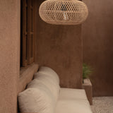 A woven natural-fiber pendant light hangs above a cream outdoor sofa in a warm, earthy patio setting with stucco walls and a small wooden side table in the foreground.