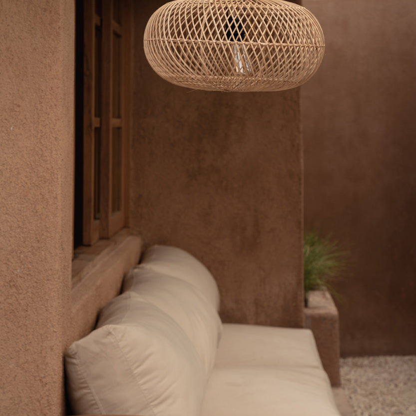 A woven natural-fiber pendant light hangs above a cream outdoor sofa in a warm, earthy patio setting with stucco walls and a small wooden side table in the foreground.