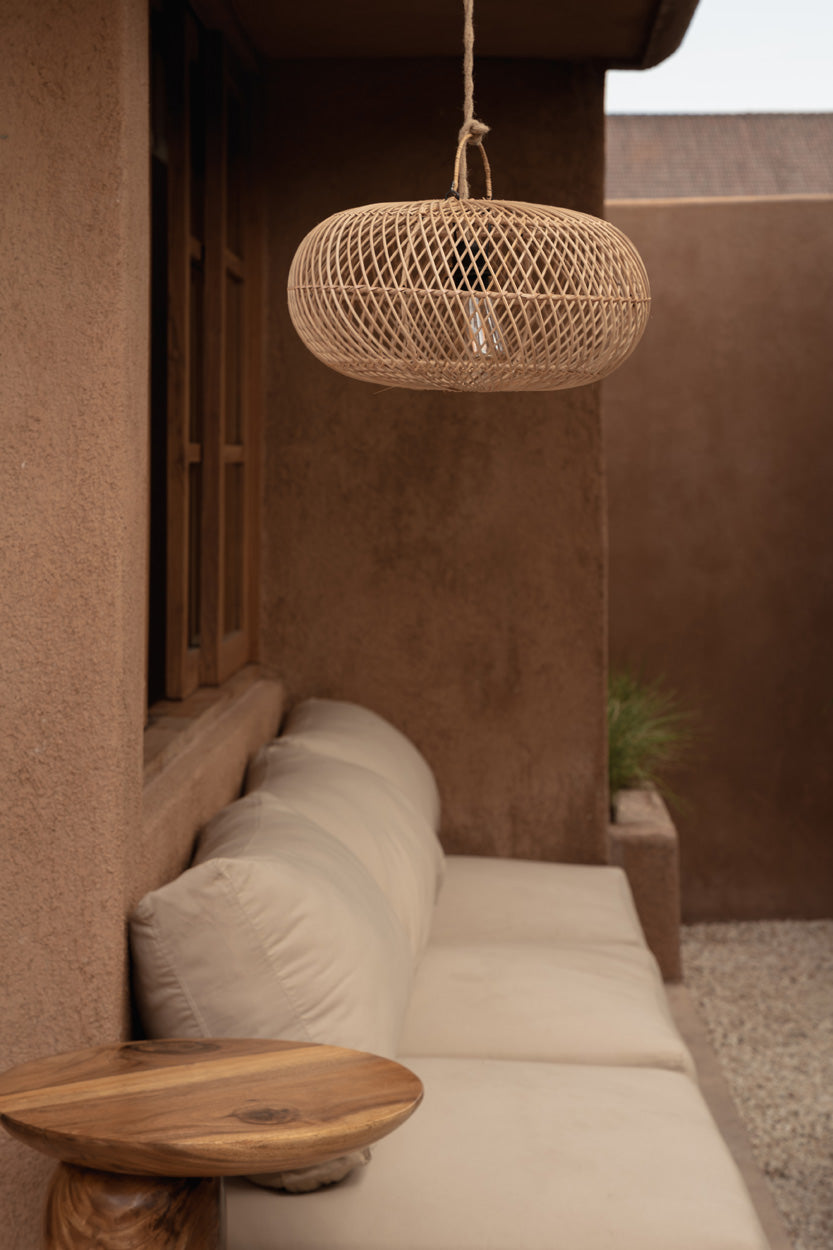 A woven natural fiber pendant light hangs above a cream outdoor sofa in a warm, earthy patio setting with stucco walls and a small wooden side table in the foreground.
