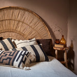 Bed with wicker headboard and decorative cushions, next to a small wooden table with a bottle and books.