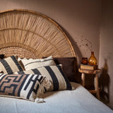 Bed with wicker headboard and decorative cushions, next to a small wooden table with a bottle and books.