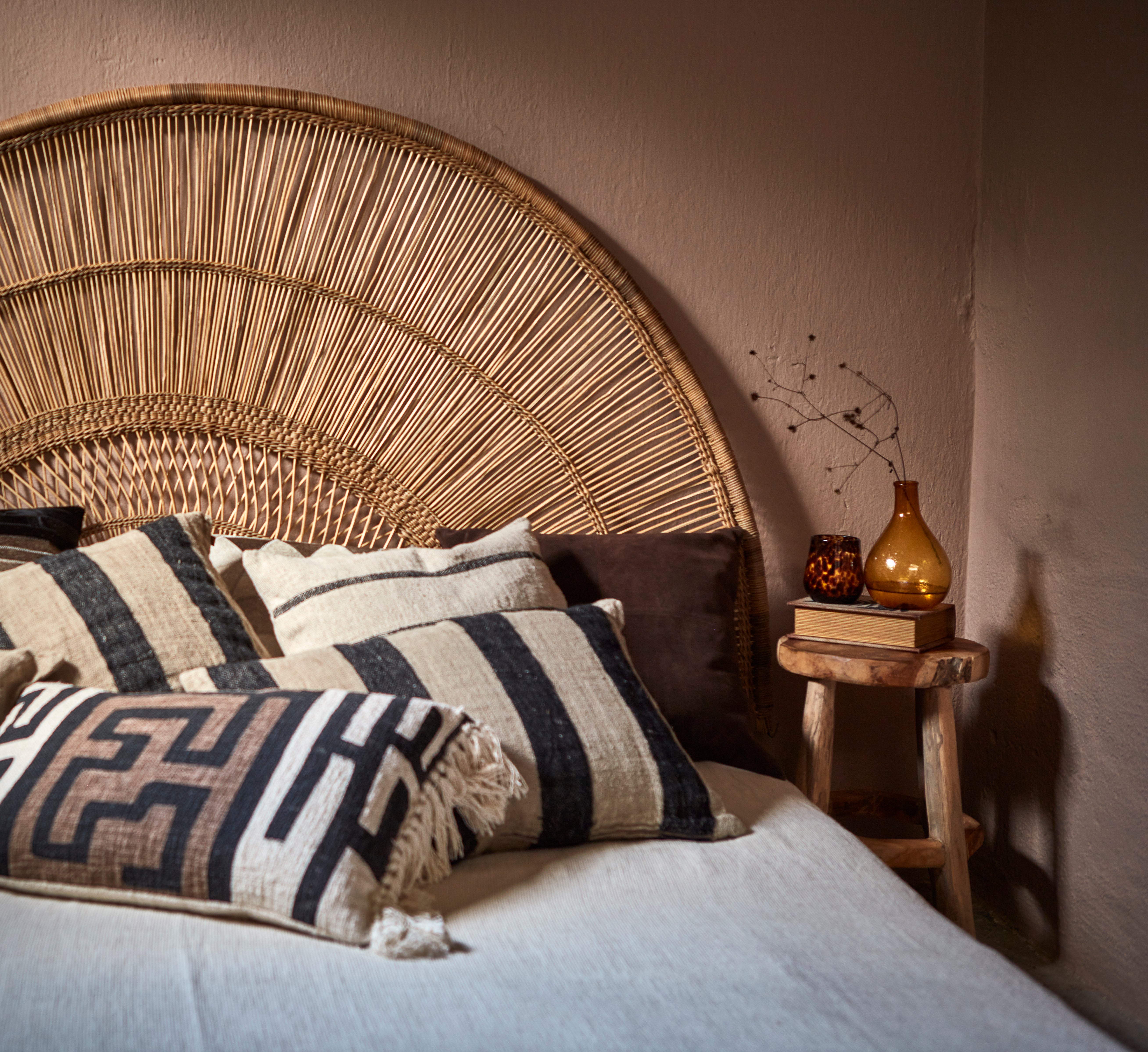 Bed with wicker headboard and decorative cushions, next to a small wooden table with a bottle and books.