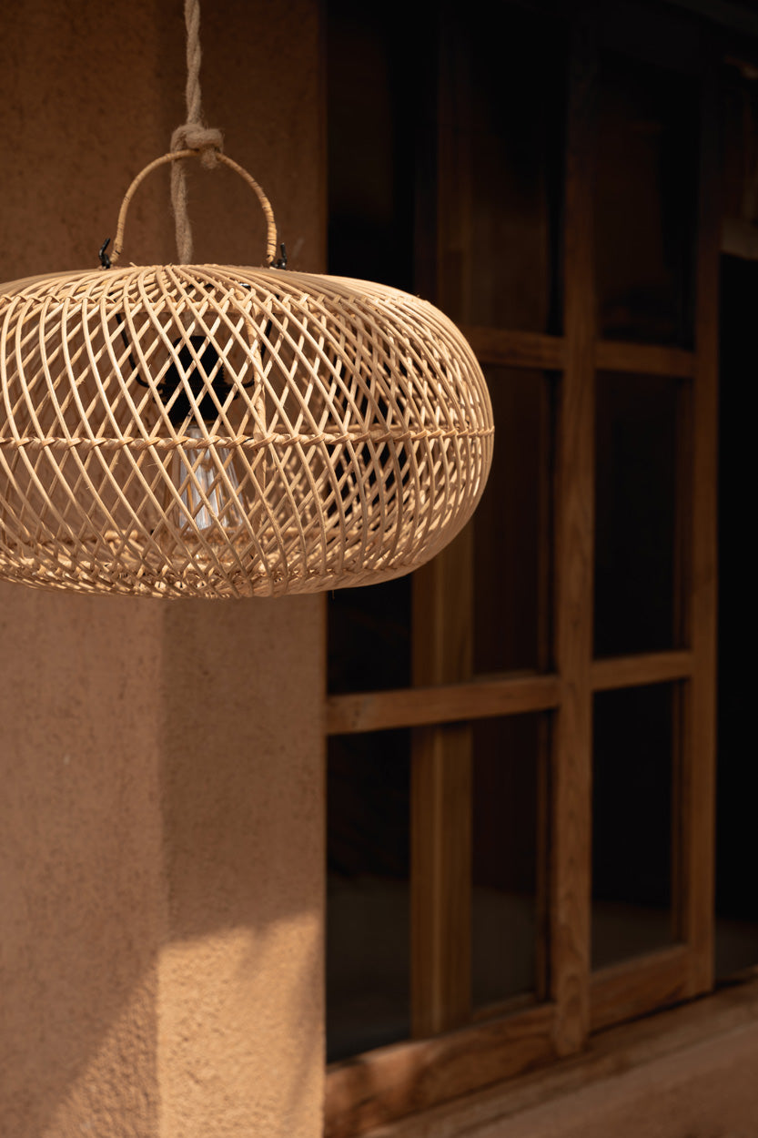 A woven natural wicker rattan  pendant light with a spherical basket shade and hanging loop, shown hanging in a warm, rustic interior setting with a wooden window and textured wall.