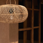 A woven natural wicker rattan  pendant light with a spherical basket shade and hanging loop, shown hanging in a warm, rustic interior setting with a wooden window and textured wall.