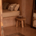 Small bedroom with a bed, wooden stool, and basket in a warm-toned room with natural lined cushions 