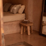 Small bedroom with a bed, wooden stool, and basket in a warm-toned room with natural lined cushions 