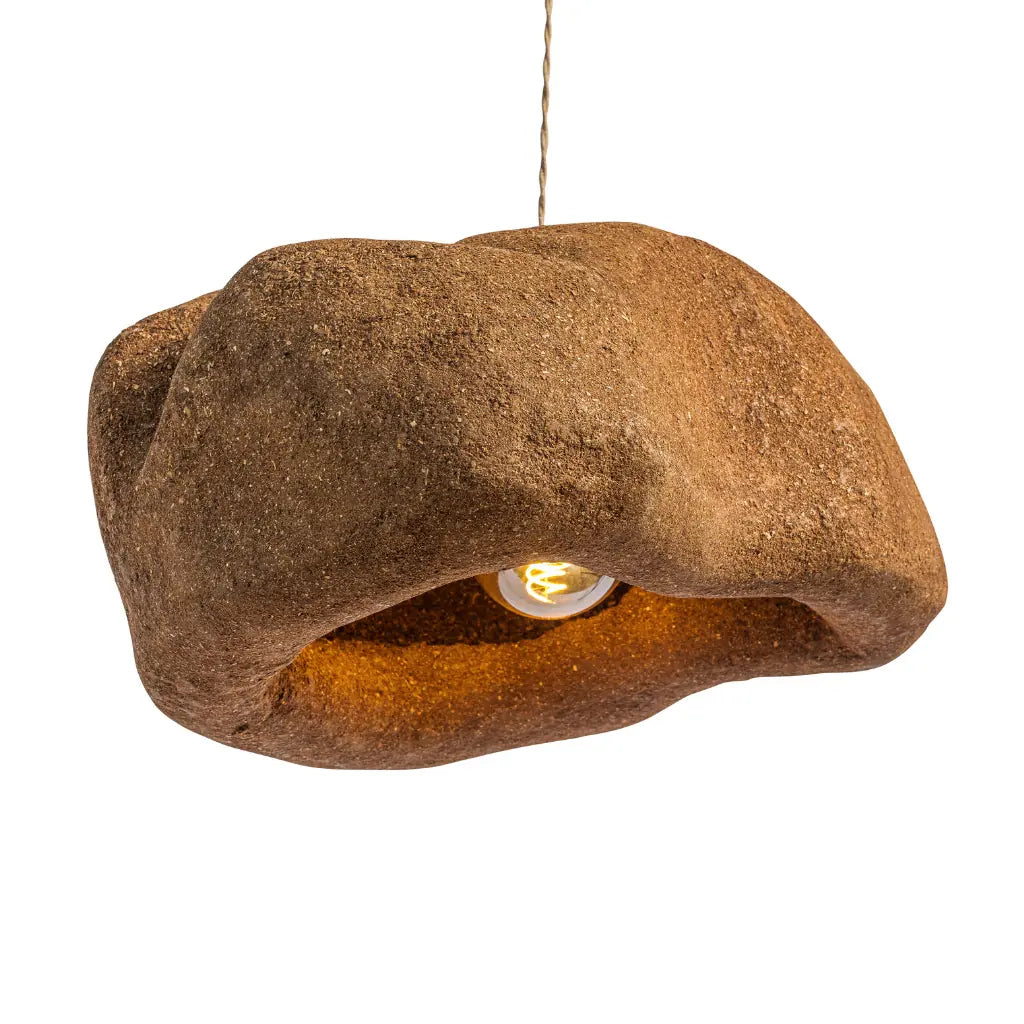 A rustic, organic-shaped pendant light fixture made from textured brown material, resembling a piece of wood or stone, with a warm glowing light bulb visible inside. It hangs from a thin cord against a solid white background.