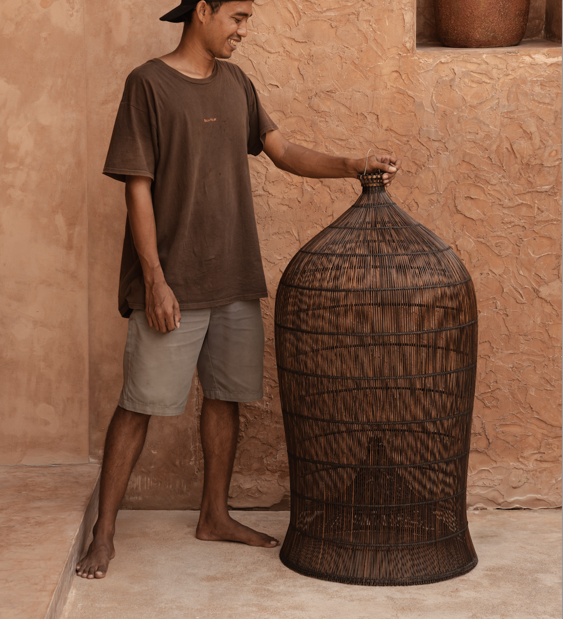 designer standing next to an extra large woven dark rattan light shade against a textured wall.