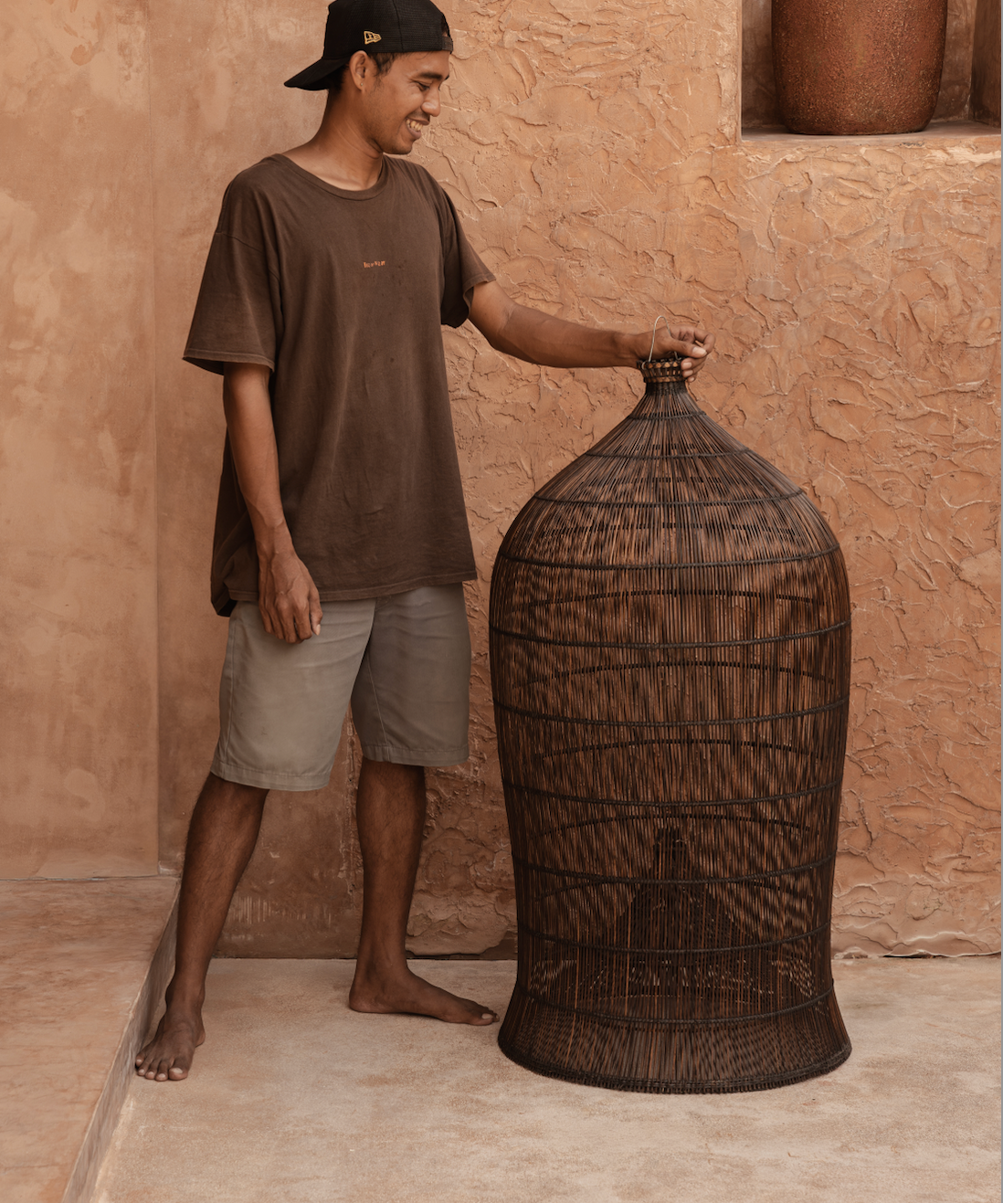 designer standing next to an extra large woven dark rattan light shade against a textured wall.