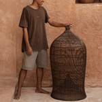 designer standing next to an extra large woven dark rattan light shade against a textured wall.
