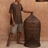 designer standing next to an extra large woven dark rattan light shade against a textured wall.