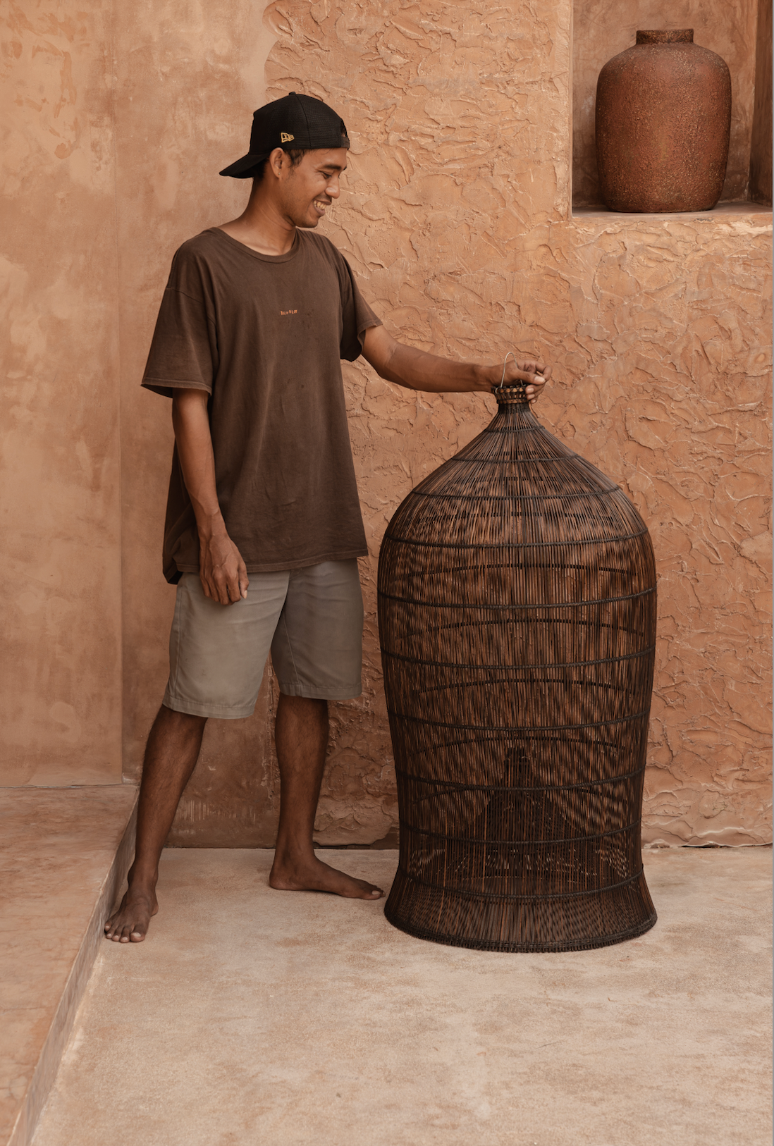 designer standing next to an extra large woven dark rattan light shade against a textured wall.