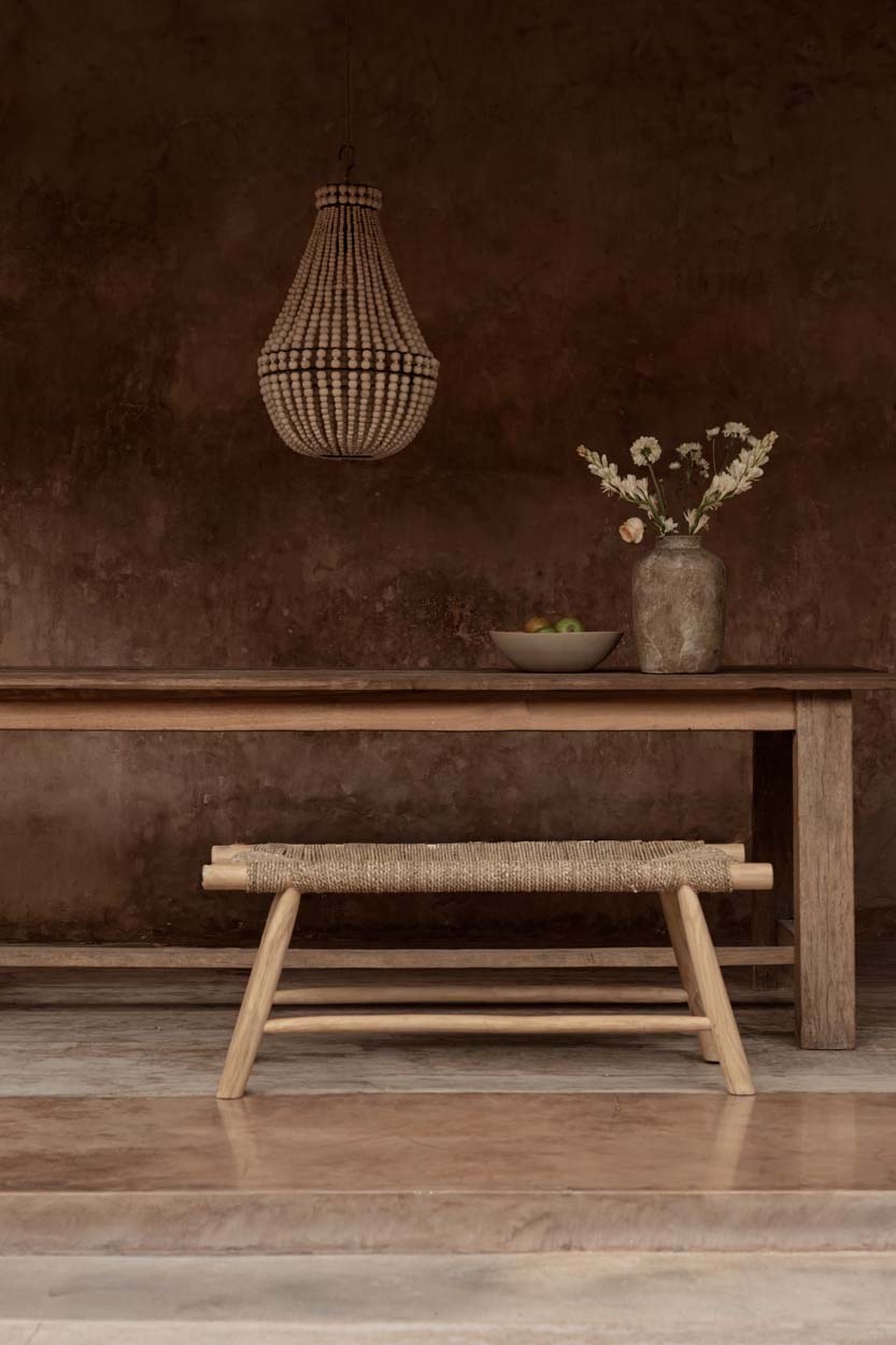 Beaded chandelier over a wooden table with a vase of flowers and a woven bench against a brown wall.