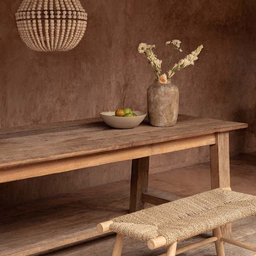 Wooden table with a vase and bowl, accompanied by a woven bench against a brown wall and wood bead chandelier