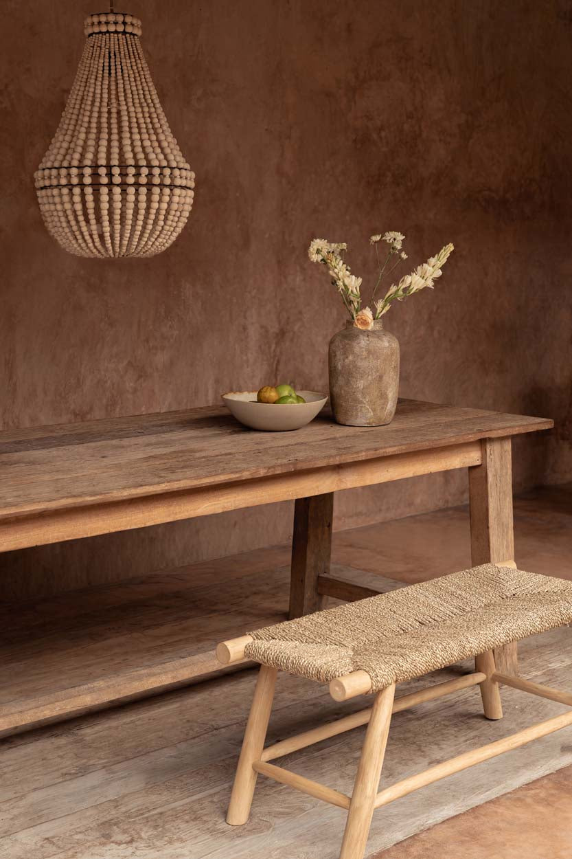 Wooden table with a vase and bowl, accompanied by a woven bench against a brown wall and wood bead chandelier