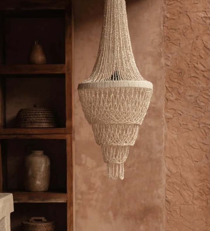 Handmade Shell chandelier  hanging against a textured wall with wooden shelves in the background.