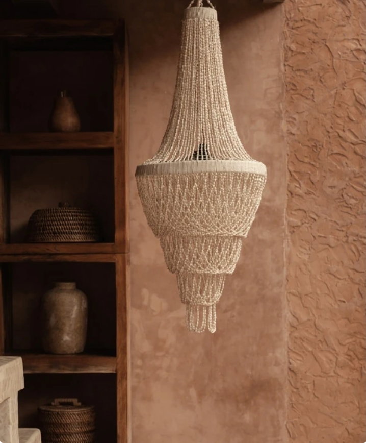 Handmade Shell chandelier  hanging against a textured wall with wooden shelves in the background.