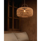 A woven rattan pendant light hanging in a rustic bedroom setting with a textured brown wall, wooden window frame, and a bed with white pillows.