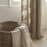 A rustic woven basket with a light beige textured throw blanket draped over the side, set in a softly lit room next to sheer beige curtains.