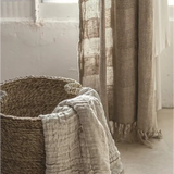 A rustic woven basket with a light beige textured throw blanket draped over the side, set in a softly lit room next to sheer beige curtains.