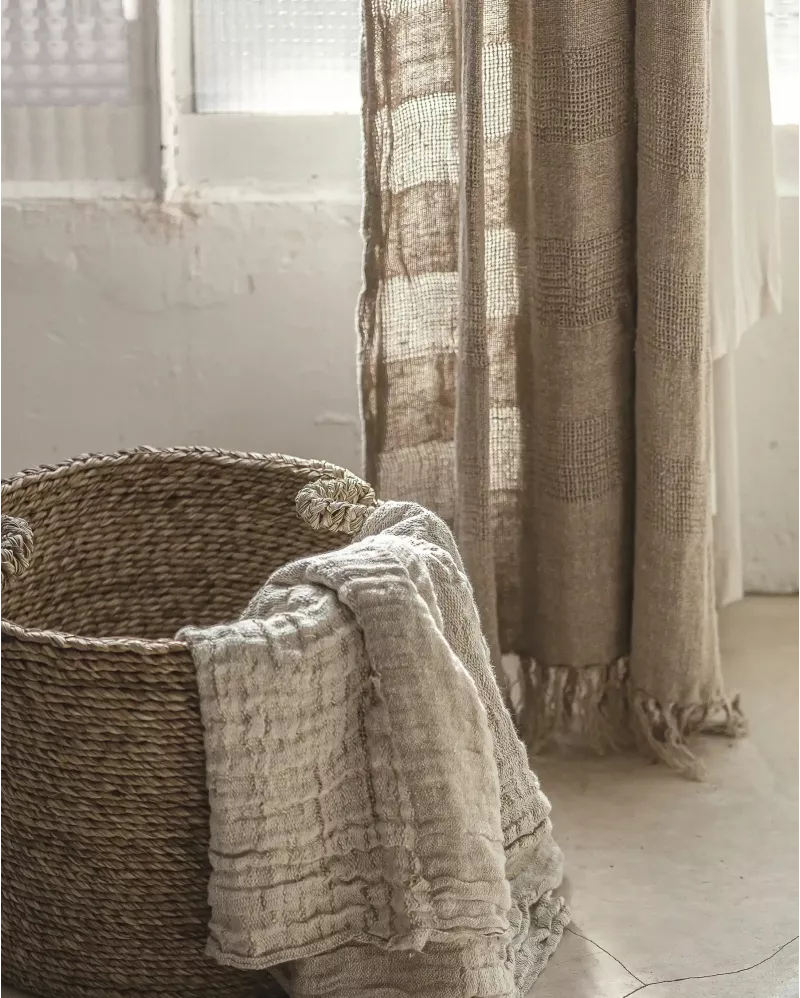 A rustic woven basket with a light beige textured throw blanket draped over the side, set in a softly lit room next to sheer beige curtains.