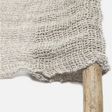 Close-up of a light beige woven textile with a rustic frayed edge draped over a wooden stick against a plain white background.