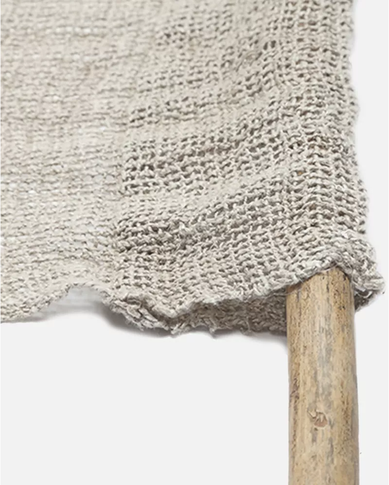 Close-up of a light beige woven textile with a rustic frayed edge draped over a wooden stick against a plain white background.