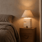Bedside table with white and linen textured lamp on a wooden nightstand next to a bed.