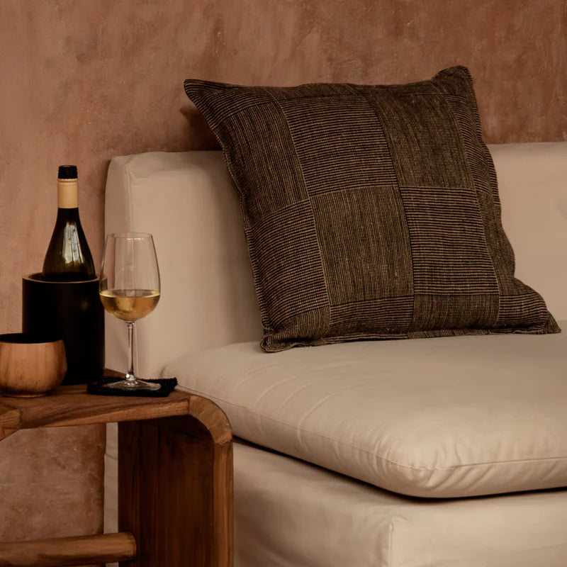 Beige sofa with a black and brown linen cushion, wine bottle, and glass on a wooden side table against a textured wall.
