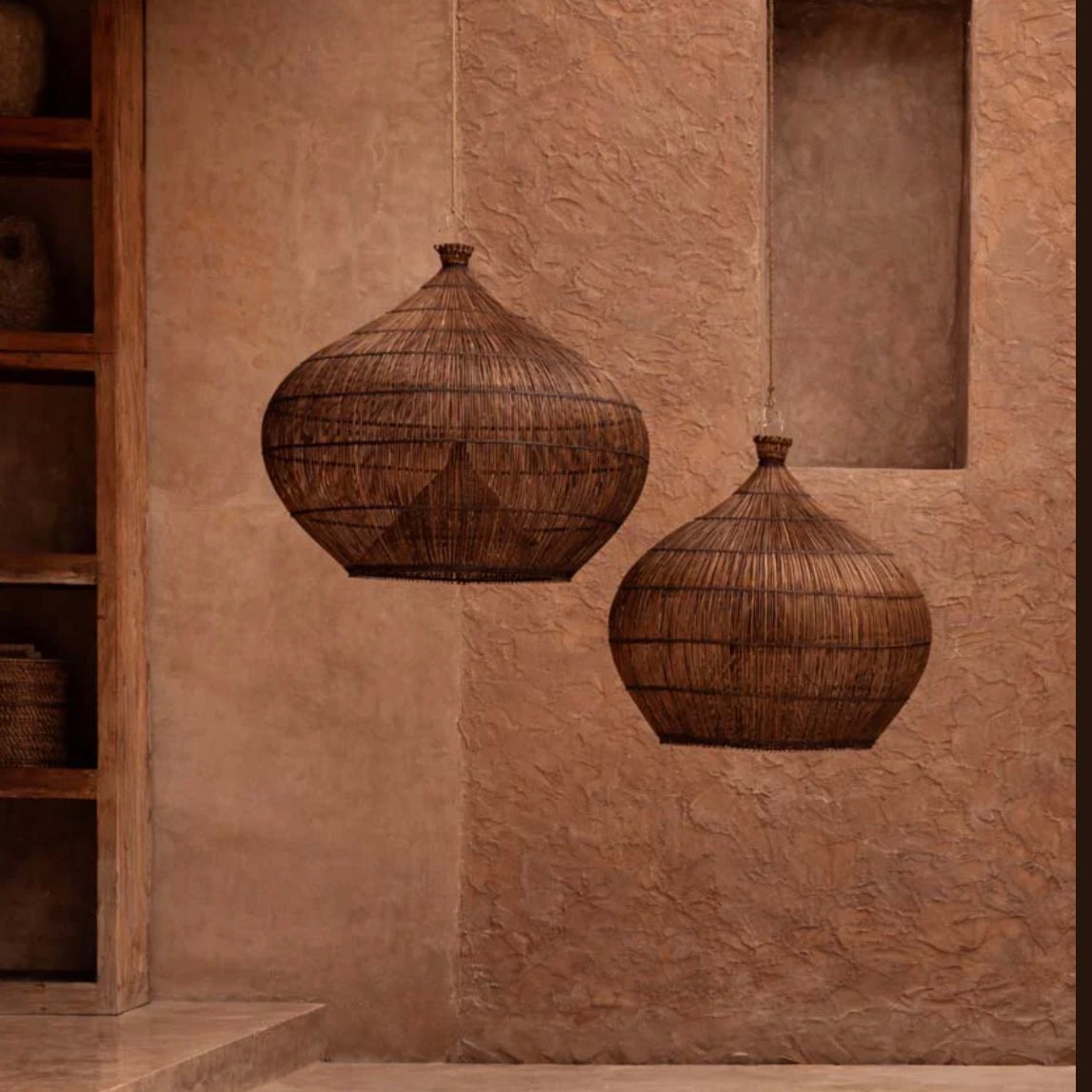 Two dark rattan pendant ceiling light shades hanging on a textured wall with a shelf and rug in the background.