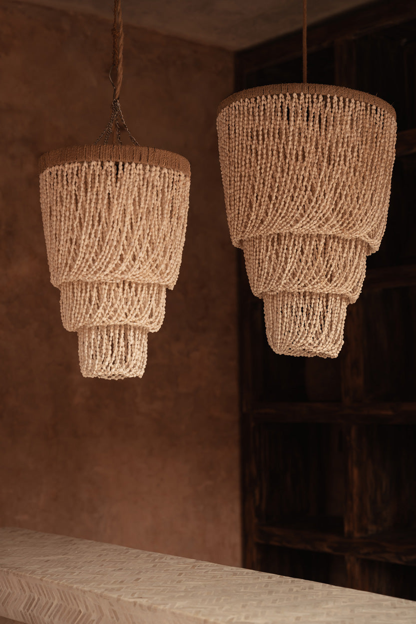 Two layered beaded shell pendant light chandeliers hanging against a brown wall.