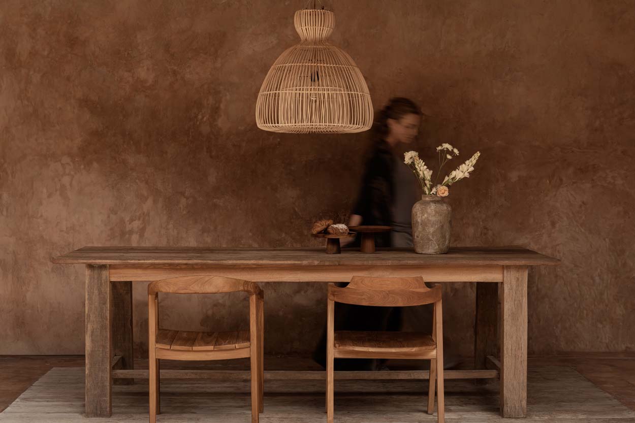 Wooden dining table with large rattan light shade with chairs against a textured brown wall.