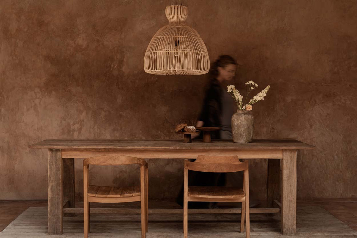 Wooden dining table with large rattan light shade with chairs against a textured brown wall.