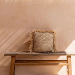 Woven raffia cushion on a wooden bench against a beige wall with soft shadows.