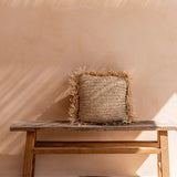 Woven raffia cushion on a wooden bench against a beige wall with soft shadows.