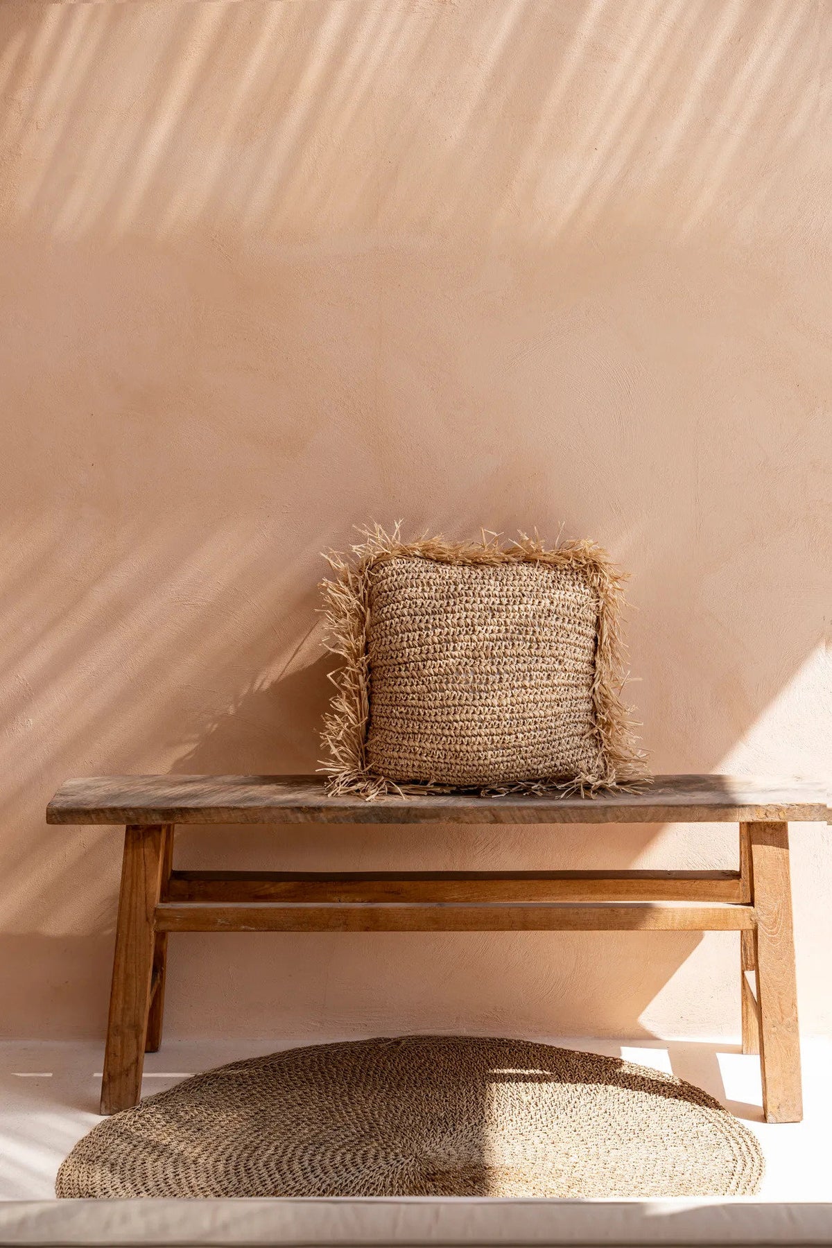 Woven raffia cushion on a wooden bench against a beige wall with soft shadows.