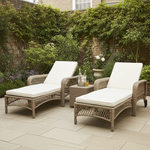 Two wicker outdoor lounge chairs with white cushions on a stone patio, surrounded by greenery.