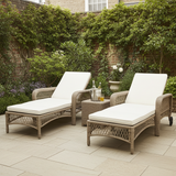 Two wicker outdoor lounge chairs with white cushions on a stone patio, surrounded by greenery.