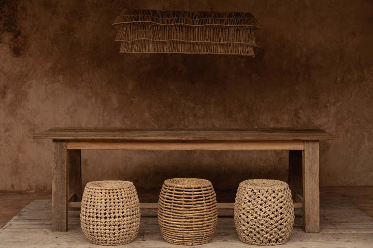 Wooden table with three woven stools against a textured brown wall and oversized rattan chandelier light